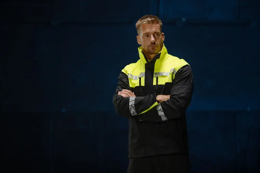 High-visibility recycled and organic work jacket worn by a worker standing with folded arms
